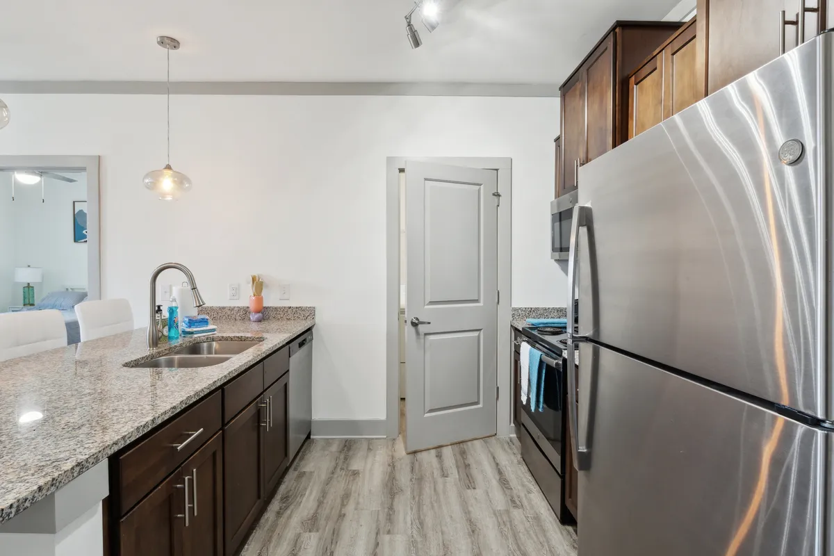 A view of a contemporary kitchen, featuring a spacious granite countertop, stainless steel appliances (including a built-in oven and refrigerator), a kitchen island with modern light fixtures overhead, and sleek cabinetry. The space is well-lit and designed for both functionality and entertaining, with clear attention to detail and organization.