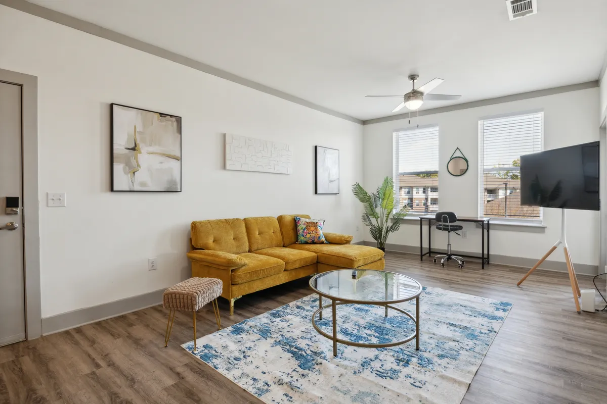 A bright and cheerful living area with a yellow sectional sofa set around a glass coffee table atop a decorative rug. Large windows flood the space with natural light, and the inviting arrangement encourages relaxation, conversation, and taking in city views. Subtle, contemporary décor and airy ambiance evoke urban sophistication.