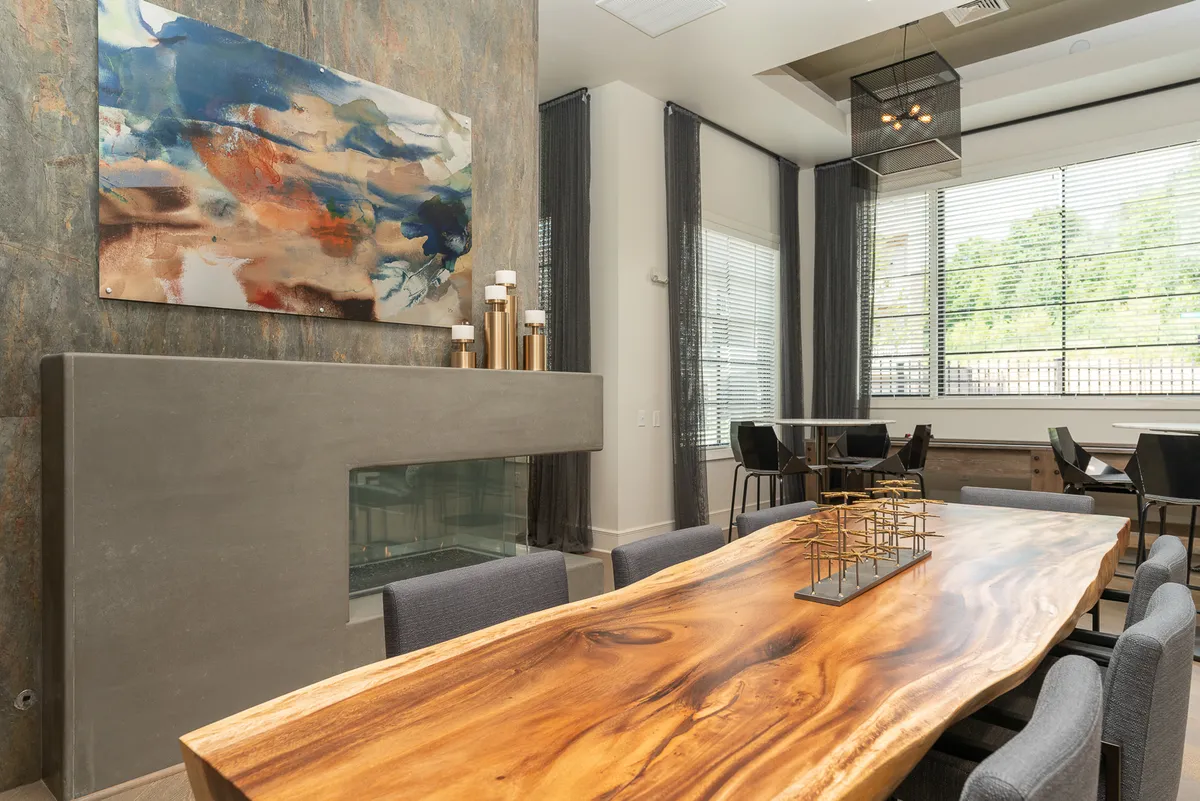 A chic dining area with a large wooden dining table, surrounded by modern chairs. Abstract wall art provides visual interest, while a concrete fireplace and black metal light fixture add warmth and style. The setting is contemporary yet welcoming, ideal for gatherings or intimate meals.