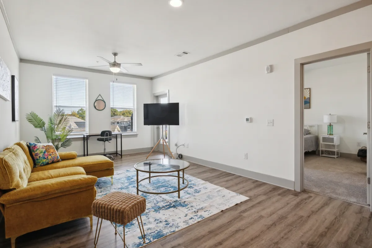A stylish living room featuring a yellow sofa, round coffee table, TV, decorative rug, and expansive windows. The space is bathed in daylight and offers a cozy environment for relaxing or entertaining, with tasteful wall art and a functional entertainment setup in view.