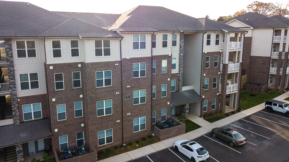 A modern brick apartment building with large windows and a covered entryway, surrounded by tidy landscaping. The setting feels both central and approachable, suggesting easy access to the surrounding city and a warm welcome home.
