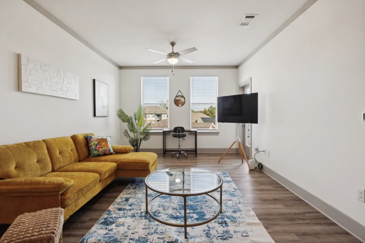 The image showcases a vibrant living area with a striking yellow couch, a modern glass coffee table, lush potted plant, sleek work desk, and a wall-mounted TV. The setting is brightly lit and thoughtfully arranged, creating a cheerful and cozy ambiance ideal for both relaxation and productivity.