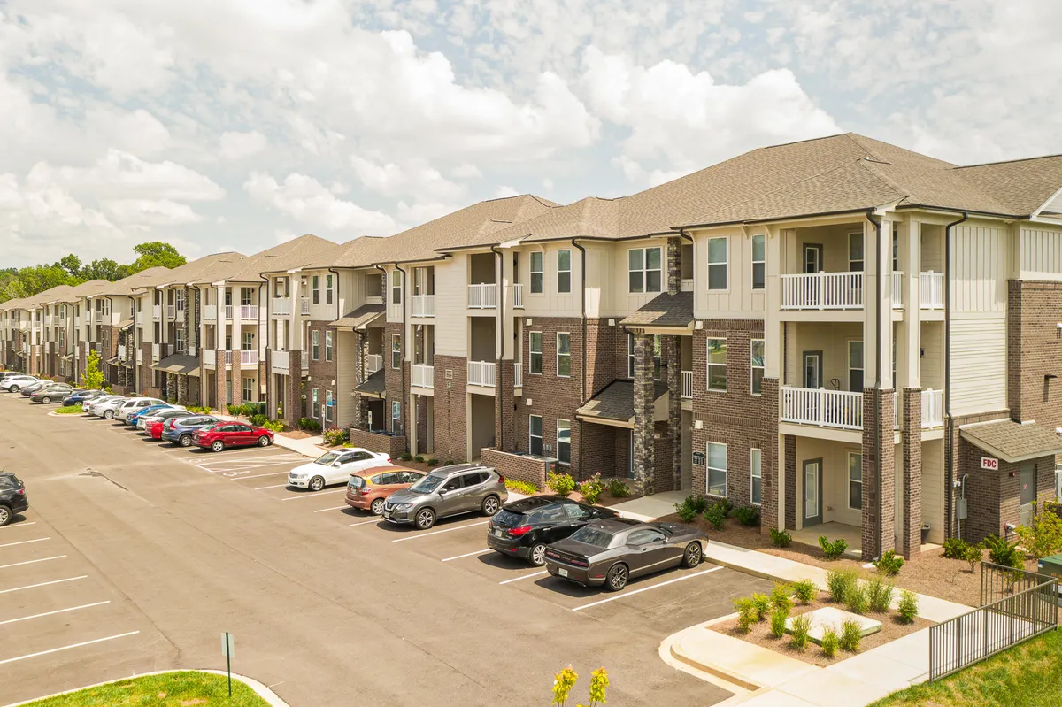 A wide-angle view of modern apartment buildings framed by attractive landscaping, with a visible parking area and open spaces where residents can gather. The welcoming exterior hints at a vibrant, urban community that connects seamlessly to the surrounding city.